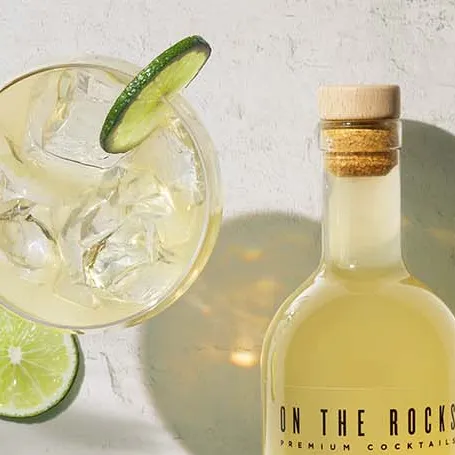 OTR cocktail bottle filled with light yellow liquid next to filled glass with ice and lime on white worktop.