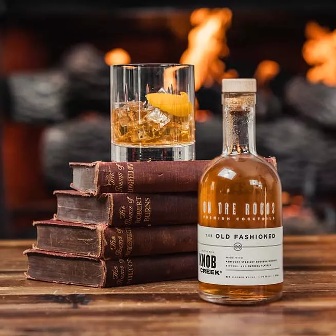 Old fashioned bottle next to a stack of old books with a filled glass on top with fireplace in background.