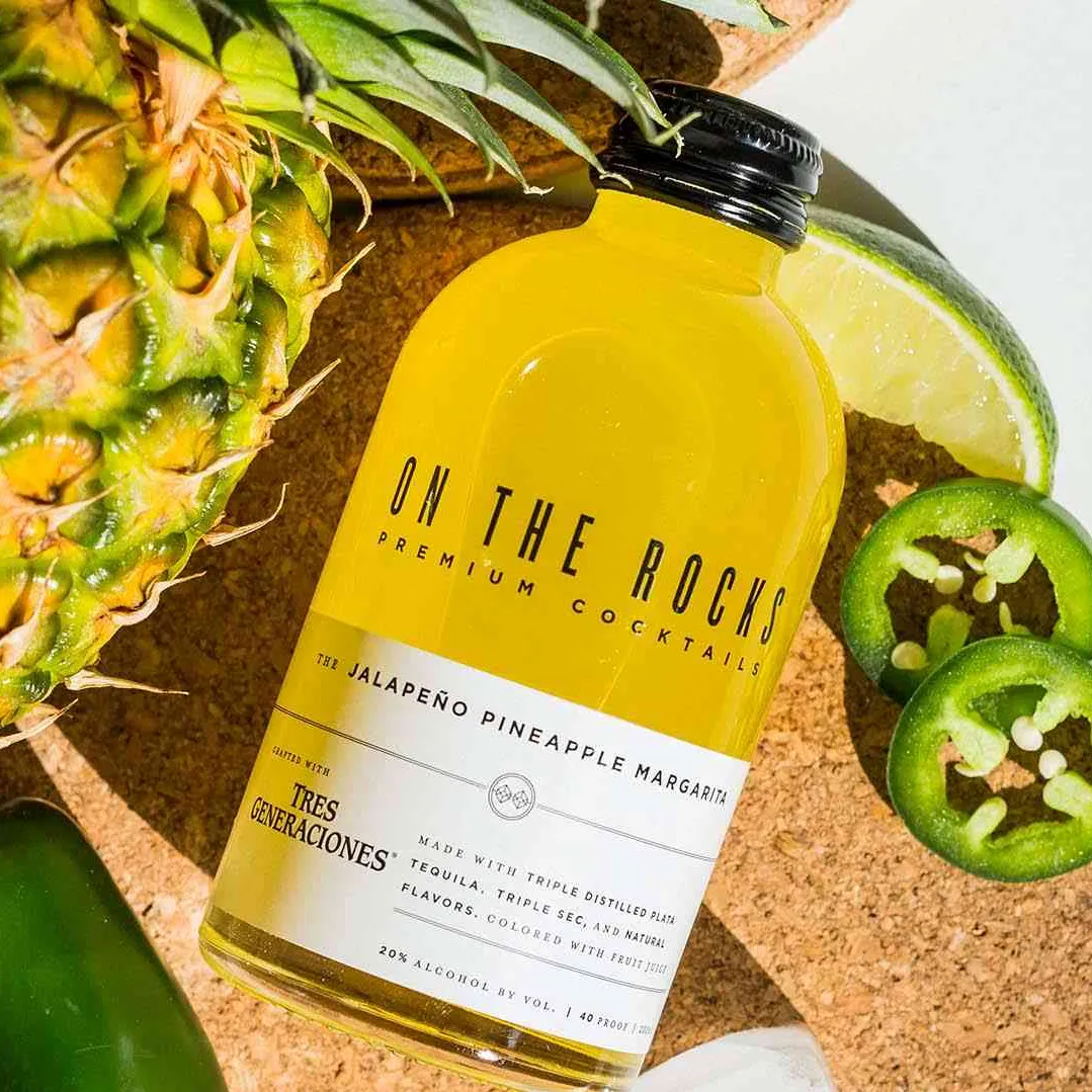 OTR Pineapple Jalapeno Margarita bottle lying next to some jalapenos, a lime and a pineapple