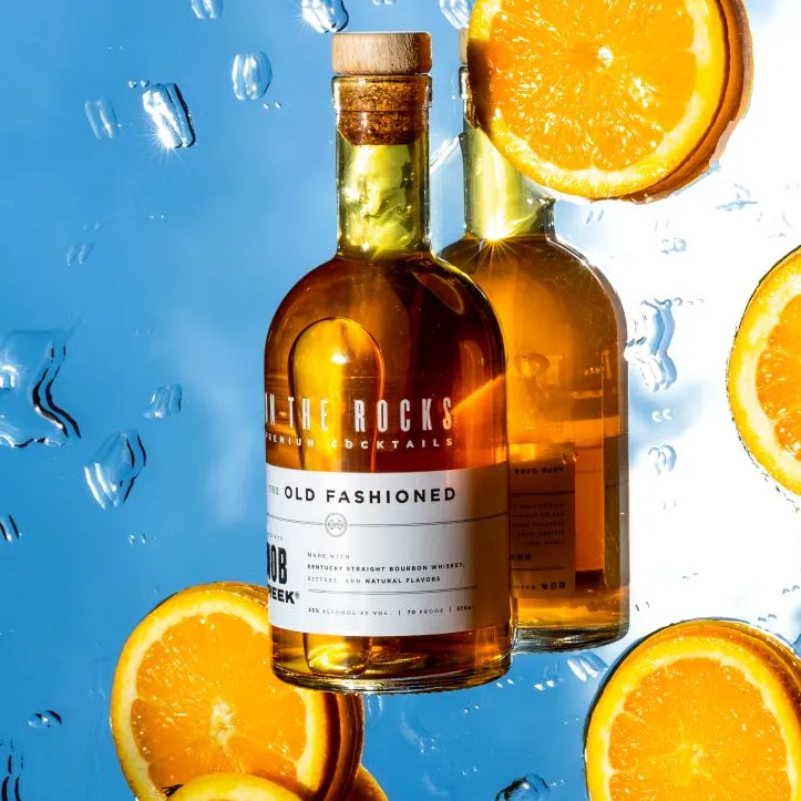 A bottle of On The Rocks Old Fashioned on a mirrored background with oranges