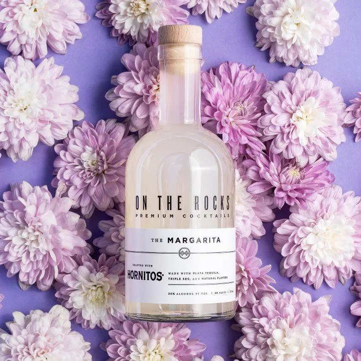 A bottle of On The Rocks Margarita on a purple background with flowers