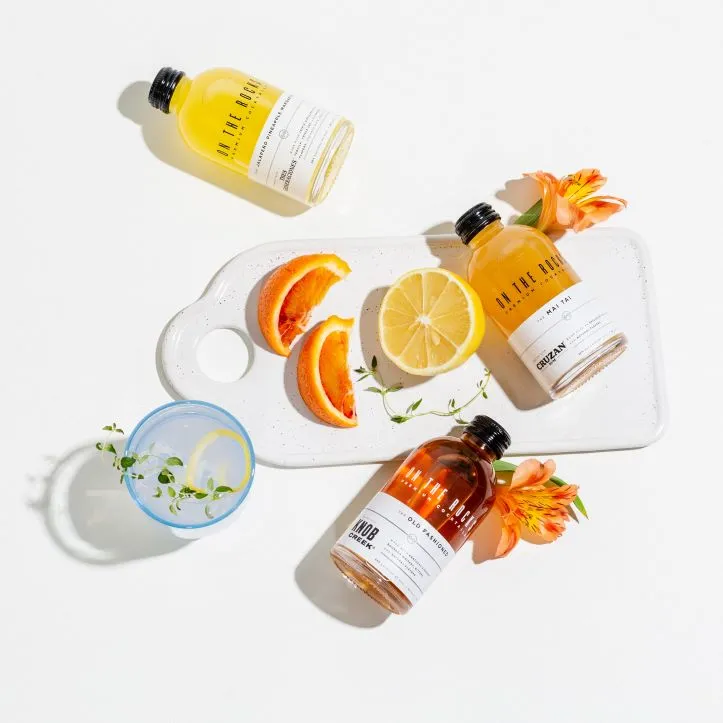 Three bottles of On The Rocks lying on a cutting board with flowers, lemons and oranges