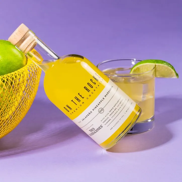 A bottle of On The Rocks Jalapeno Pineapple Margarita between a fruit basket and a cocktail glass
