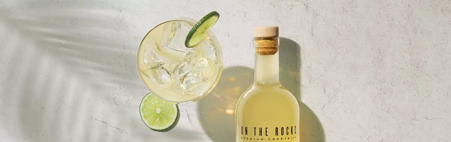 OTR cocktail bottle filled with light yellow liquid next to filled glass with ice and lime on white worktop.