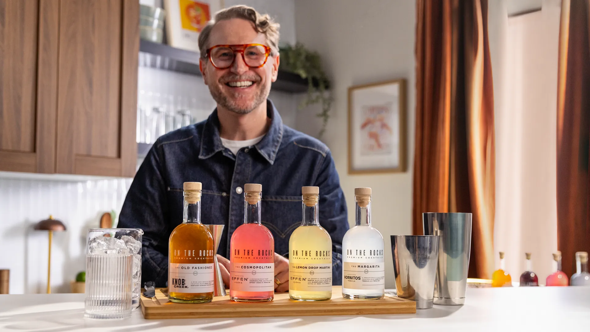 A wooden tray displays four cocktail bottles and a glass filled with ice