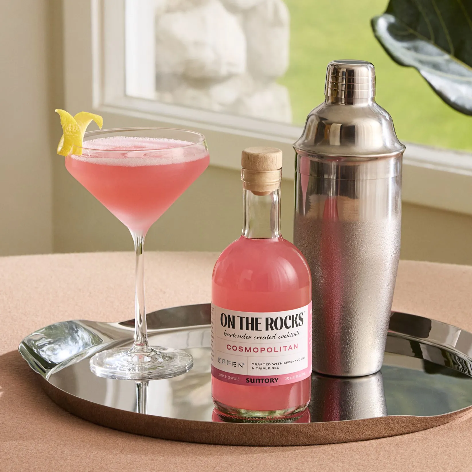 A pink Cosmopolitan cocktail in a coupe glass with a lemon twist, a matching bottled drink, and a metal shaker on a silver tray.