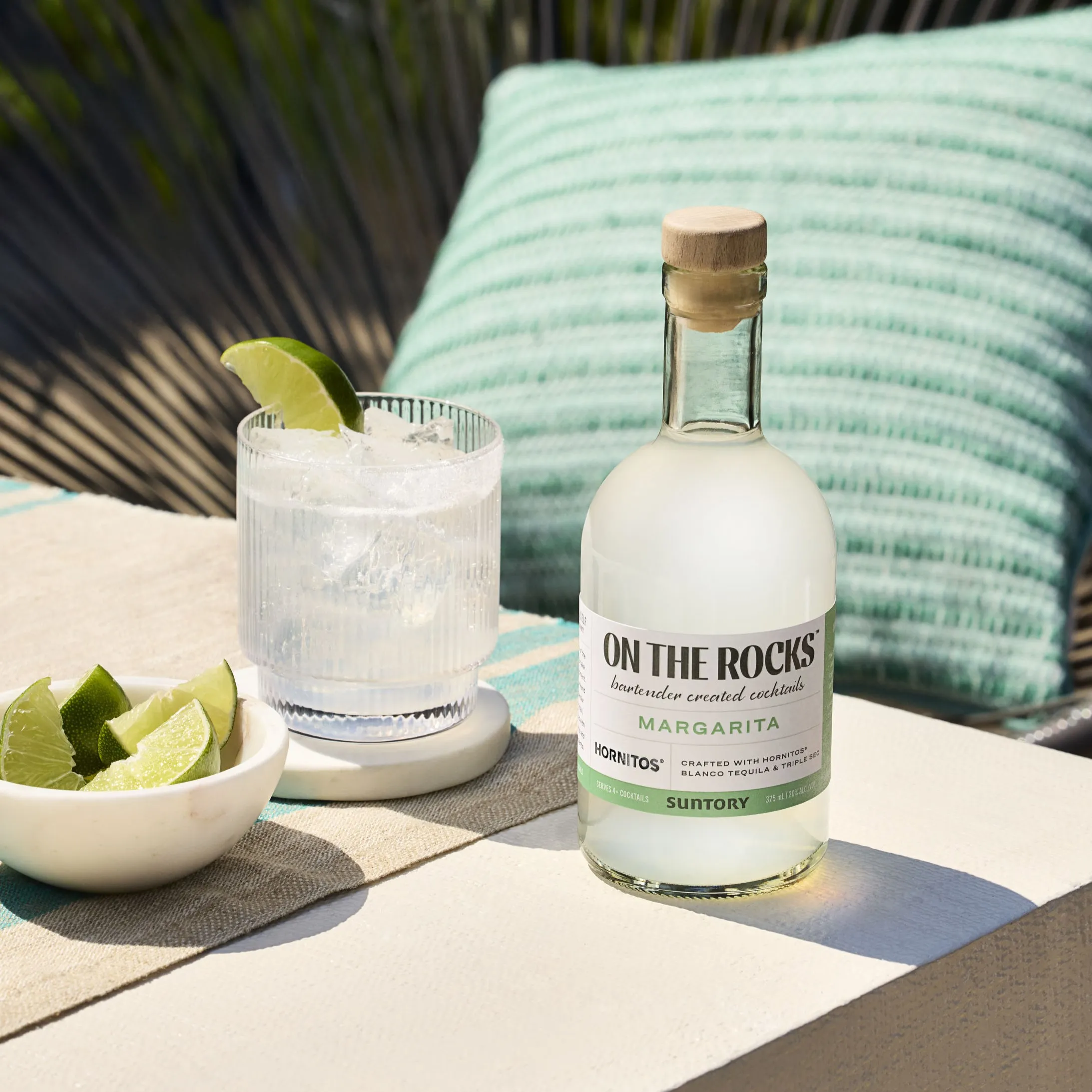 A bottle of "On The Rocks margarita" cocktail stands next to a glass with ice and lime wedges.