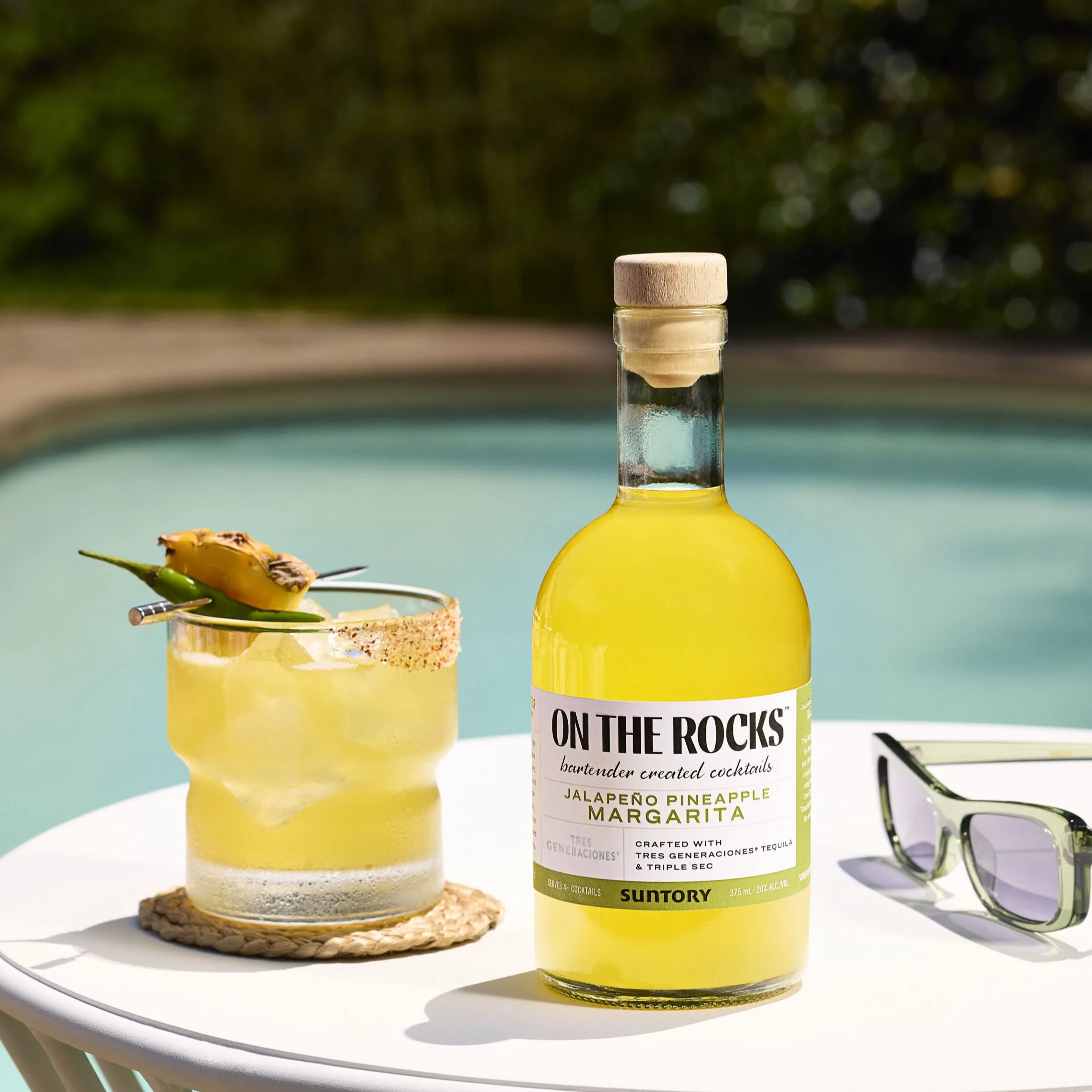 Bottle of "On The Rocks Jalapeño Pineapple Margarita" next to a cocktail glass garnished with pineapple by a pool.