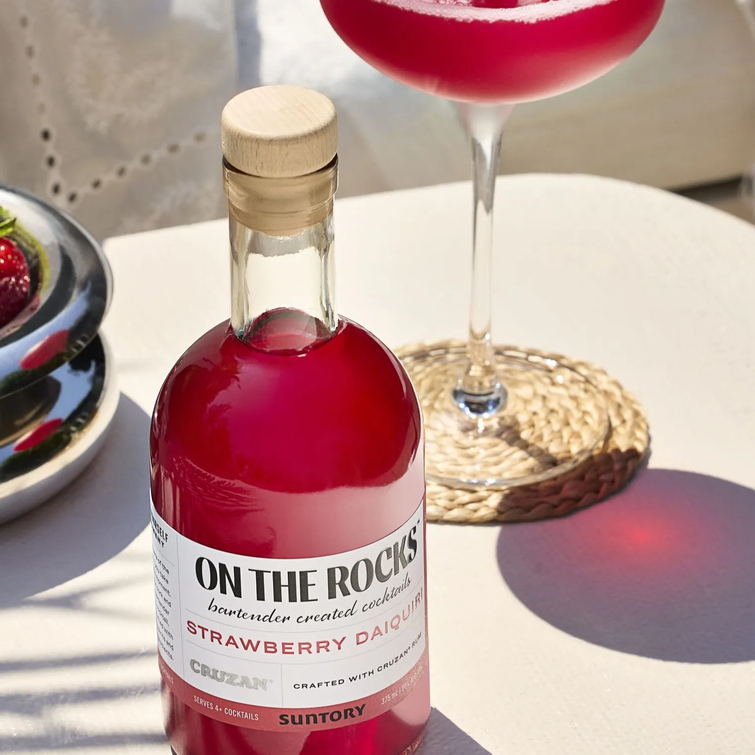 A bottle of On The Rocks Strawberry Daiquiri cocktail and a matching cocktail glass on a table, with sunlight casting shadows, creating a refreshing and vibrant mood.