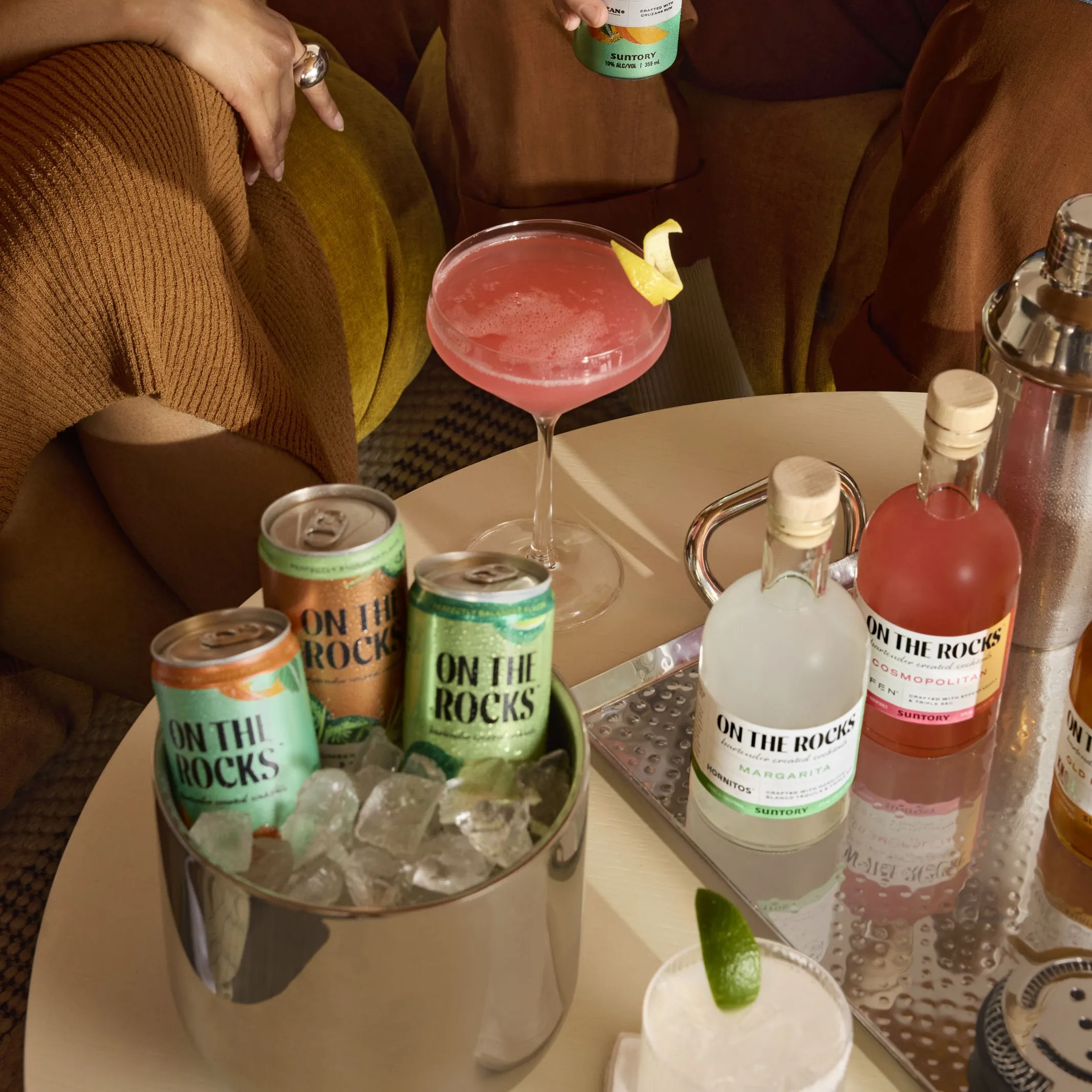 A table with assorted "On the Rocks" drinks: a pink cocktail with a lemon twist, cans in ice, and bottled drinks. Cozy, relaxed gathering vibe.