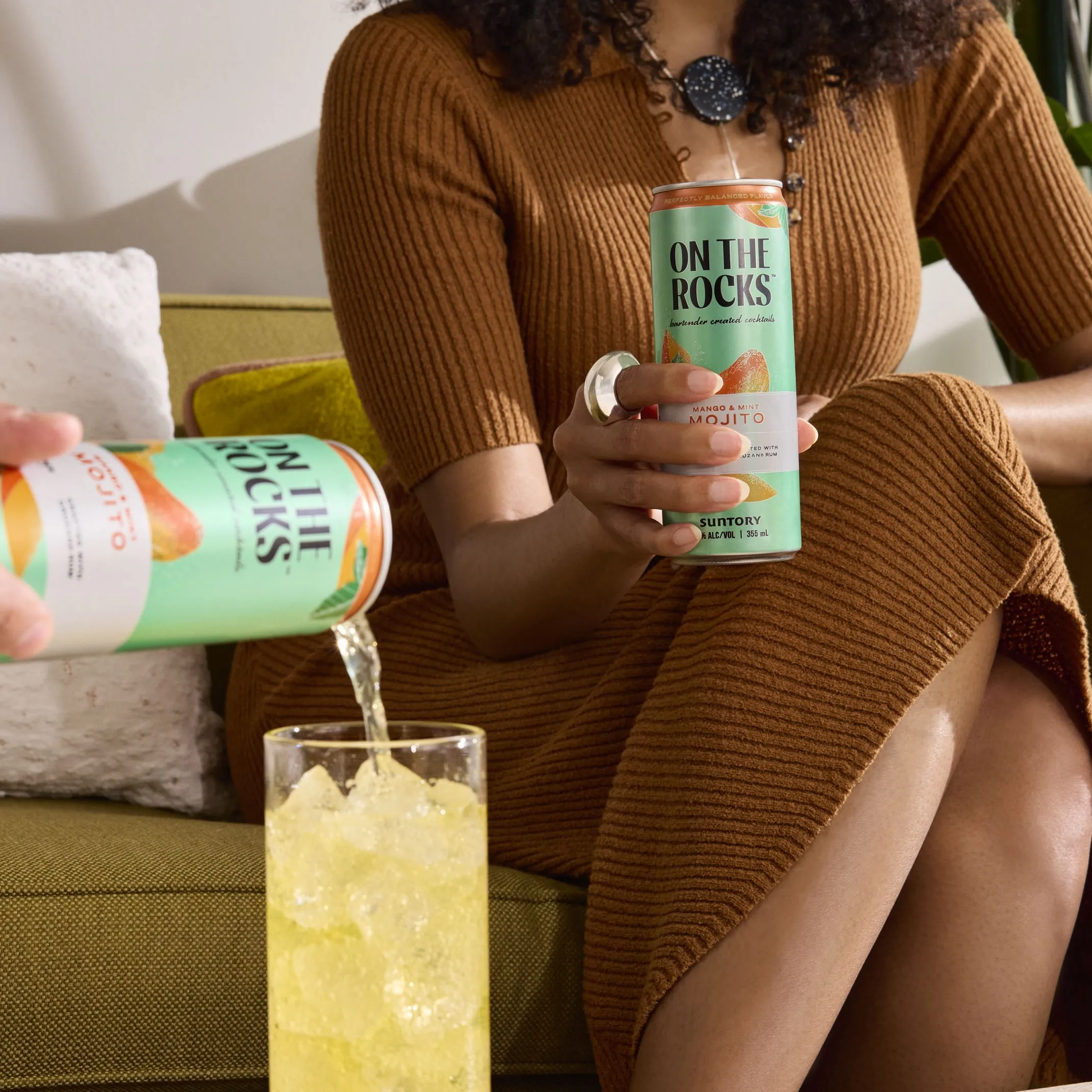 A woman in a brown dress holds a canned mojito while another can is poured into a glass with ice on a couch.
