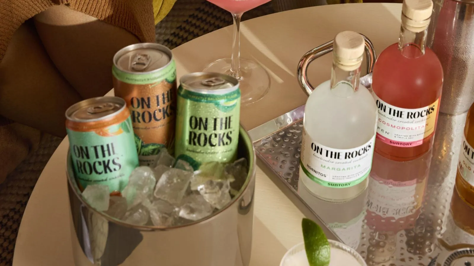 A stylish drink setup featuring colorful cans and bottles of "On The Rocks" cocktails, surrounded by ice and garnished with lime.