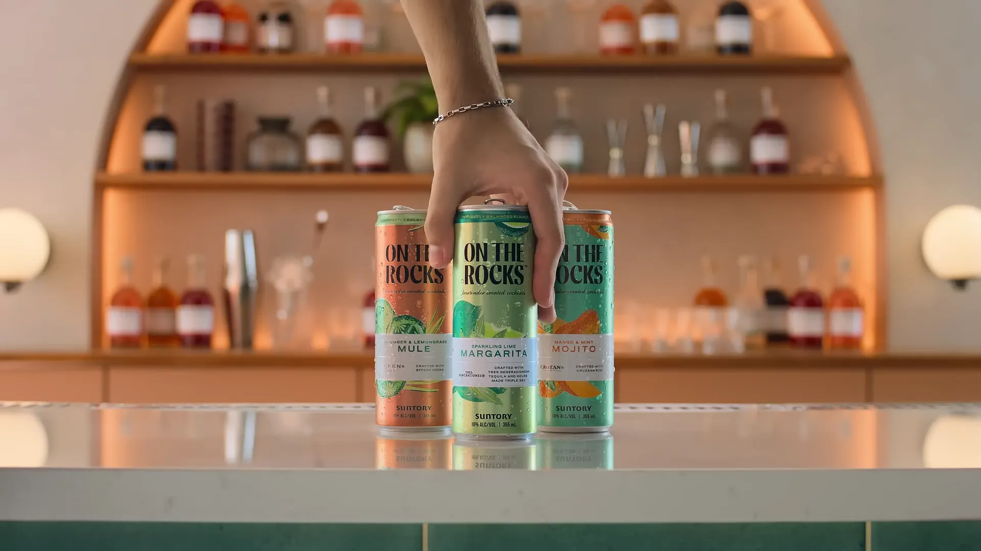 A hand holds three colorful canned cocktails labeled Mule, Margarita, and Mojito, set against a stylish bar backdrop.