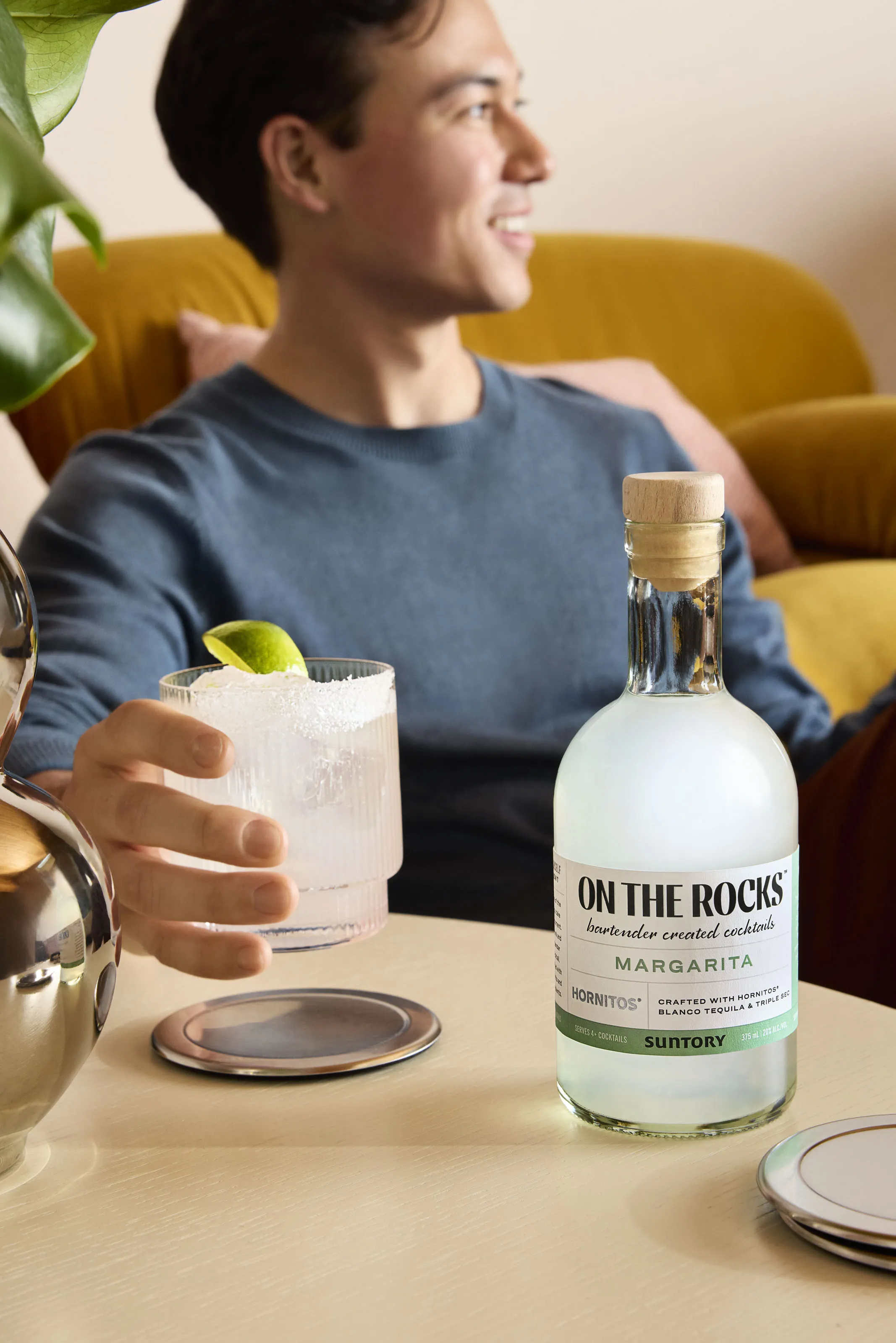 A hand holds a cocktail glass with salt, next to a bottle of "On The Rocks" Margarita, on a stylish coffee table.