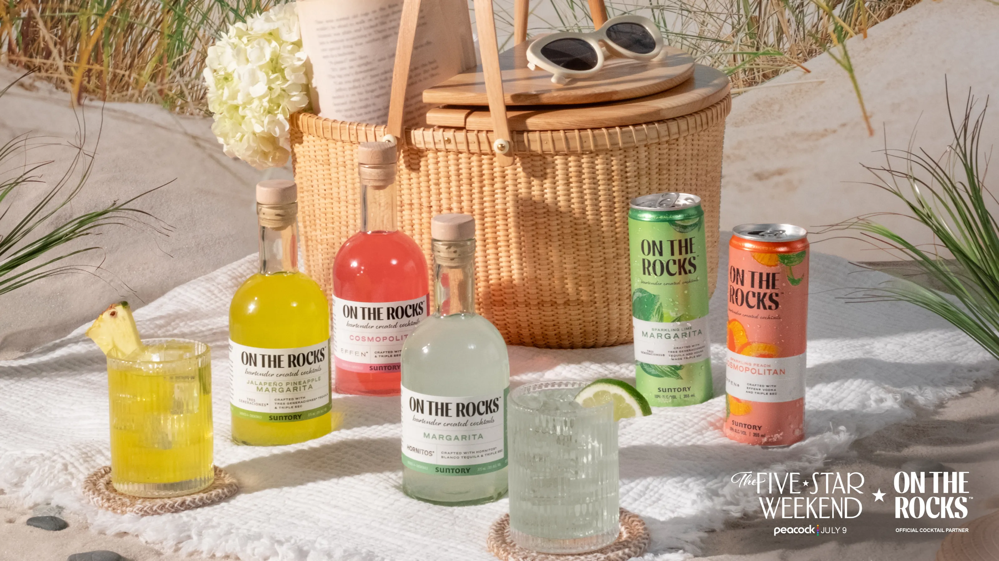 A beach picnic setup with bottles and cans of "On The Rocks" cocktails, a wicker picnic basket with sunglasses and books, and two finished cocktails on coasters.